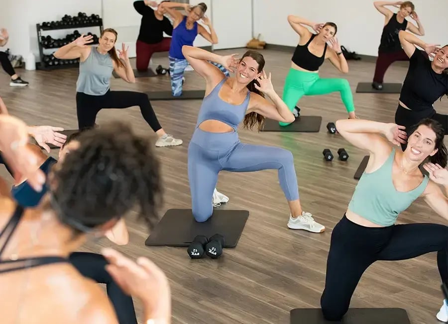 A group fitness class with people performing exercises on mats in a bright studio. Participants wear various activewear, and some hold their hands behind their heads while lunging. Light wooden floor and gym equipment are visible in the background.
