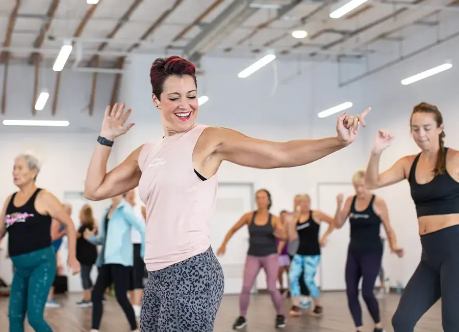 Jazzercise customer doing an energetic dance workout class with other customers smiling and moving enthusiastically in a bright, modern studio.