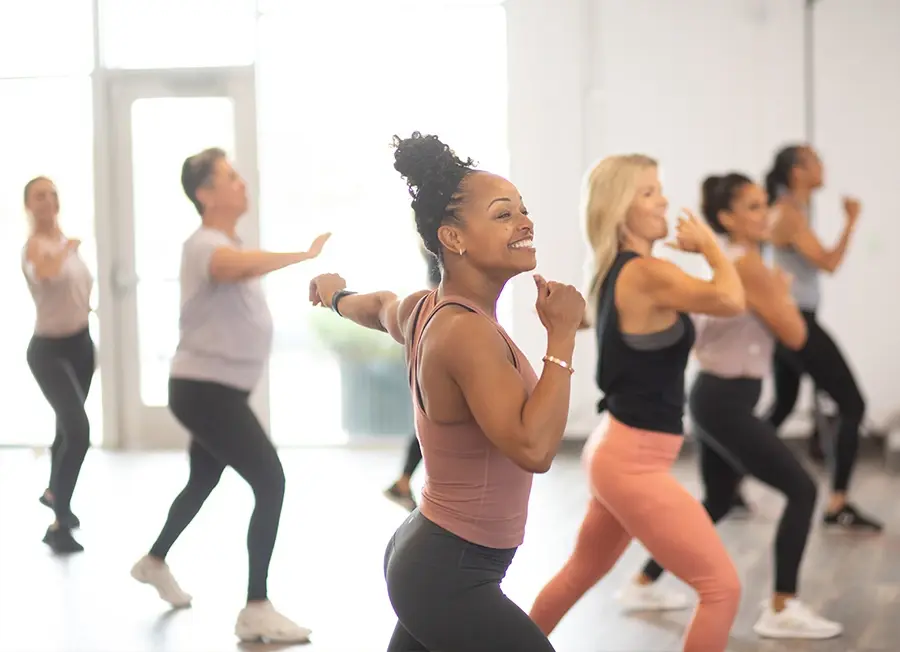 A group of women participates in a fitness class, performing lunges in a bright studio. They are wearing various workout outfits and appear focused and energetic. The room has large windows letting in natural light.